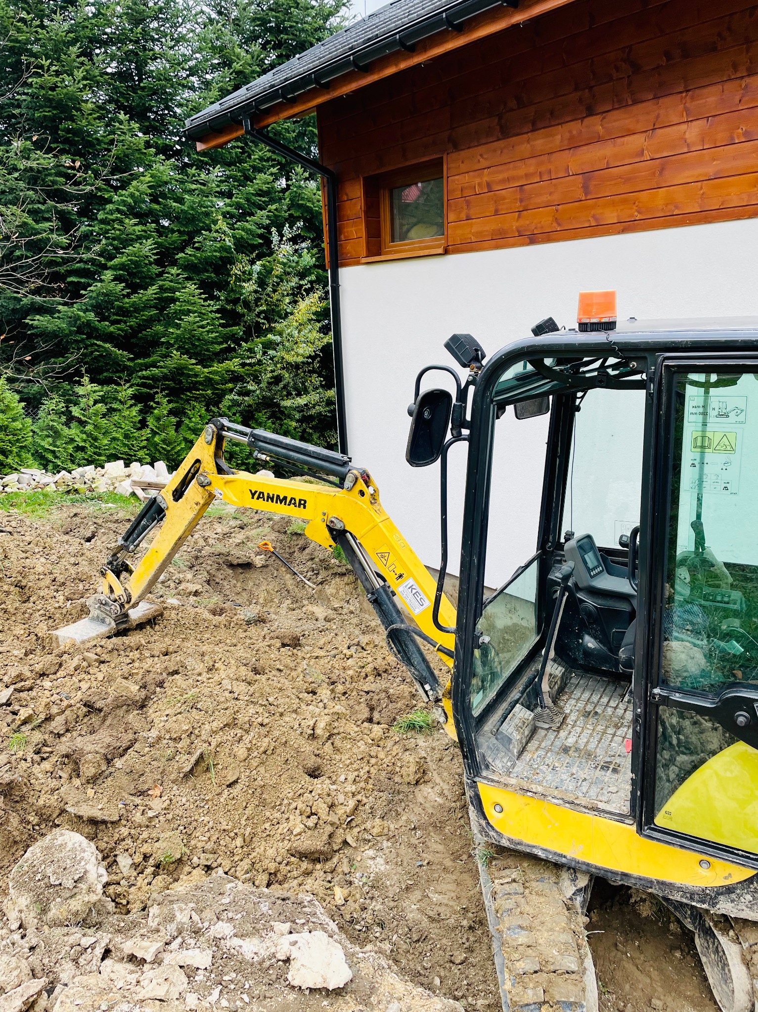 Żółta minikoparka Yanmar w trakcie prac ziemnych przy budynku z drewnianą elewacją, widoczne wykopane fragmenty ziemi i kamieni.
