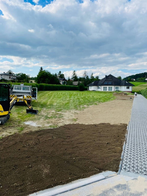 Przygotowanie terenu pod trawnik: wyrównana, ciemna ziemia, piasek, zielony trawnik, mini koparka i dom w tle.