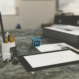 Biurko grafika z tabletem, kubkiem z przyborami, kostką z logo Photoshop i szkicami na tle okna.