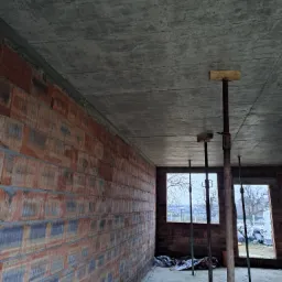 Wnętrze surowego domu w trakcie budowy: ściany z czerwonej cegły, betonowy sufit podparty metalowymi stemplami, widok przez okna na szare, zimowe otoczenie.