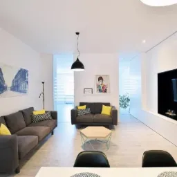 Jasne, nowocześnie urządzone wnętrze salonu z szarą sofą, obrazami z motywami miejskimi na ścianie i minimalistycznym oświetleniem.