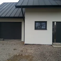 Nowoczesny dom jednorodzinny z szarą bramą garażową segmentową, drzwiami zewnętrznymi w kolorze antracytowym i czarnym dachem z blachy na rąbek stojący, teren wokół domu wysypany żwirem.