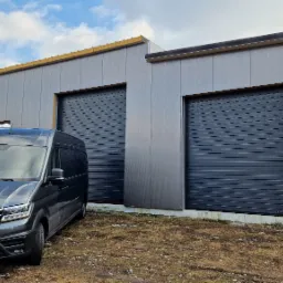 Szara hala przemysłowa z dwiema zamkniętymi, rolowanymi bramami w kolorze grafitowym i zaparkowanym przed nią ciemnym vanem Volkswagen Crafter.