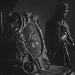 Czarno-białe ujęcie sakralnej rzeźby Matki Boskiej z aureolą, stojącej obok zdobionej, drewnianej tablicy z inskrypcjami w kościelnym wnętrzu.