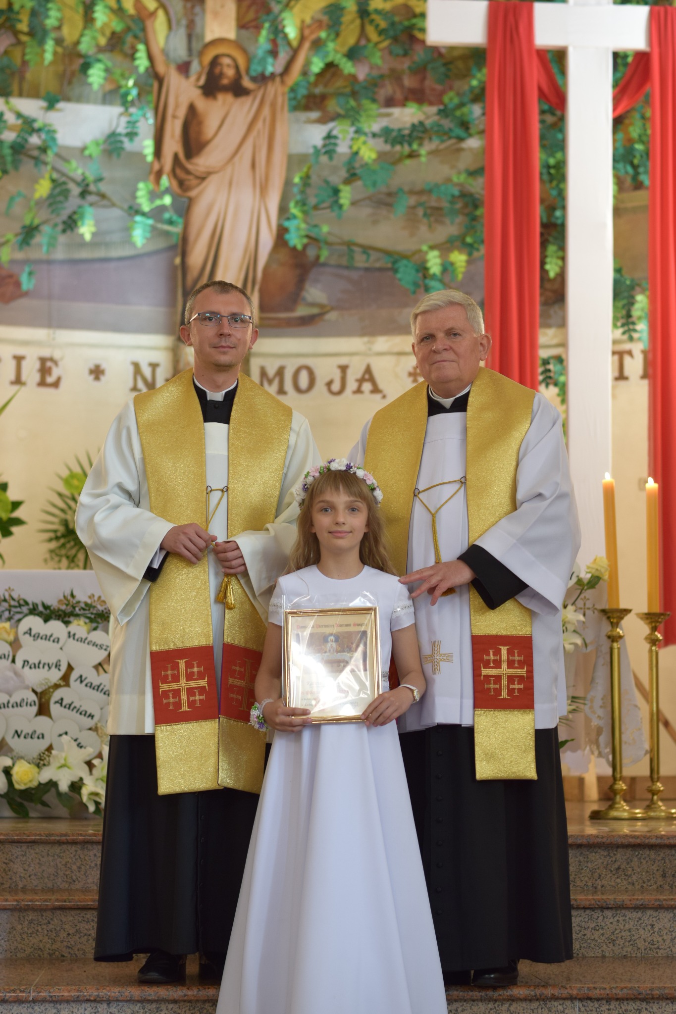 Dziewczynka w białej sukience i wianku na głowie, trzymająca oprawiony obrazek, pozuje do zdjęcia z dwoma księżmi w kościele podczas uroczystości Pierwszej Komunii Świętej.