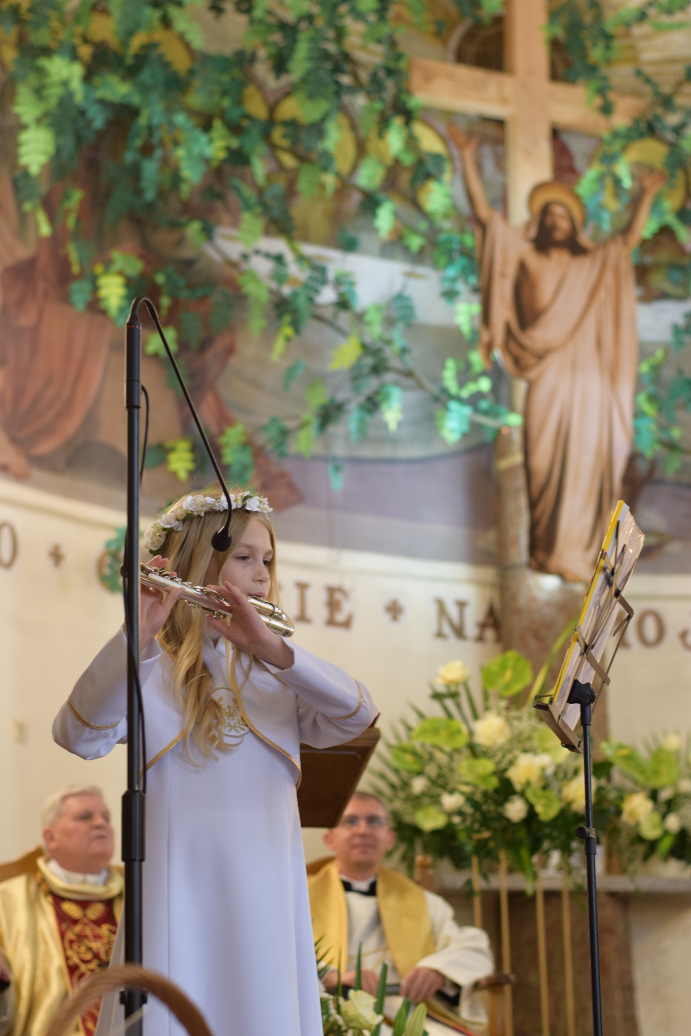 Dziewczynka w białej albie i wianku gra na flecie podczas uroczystości w kościele, w tle widoczni księża i dekoracja roślinna.