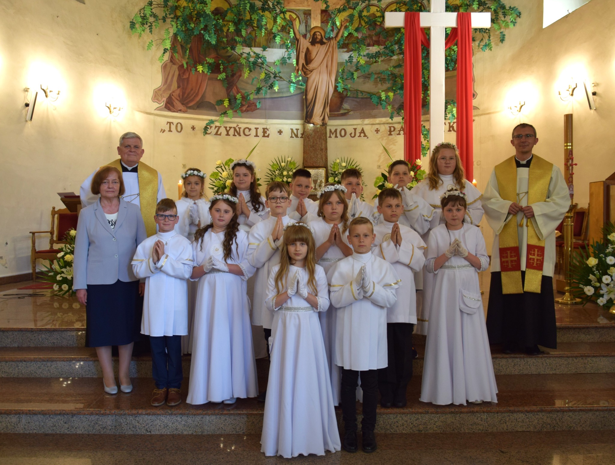 Grupa dzieci w białych strojach komunijnych z rodzicami i księżmi pozuje do zdjęcia w kościele na tle ołtarza z krzyżem i dekoracjami roślinnymi.