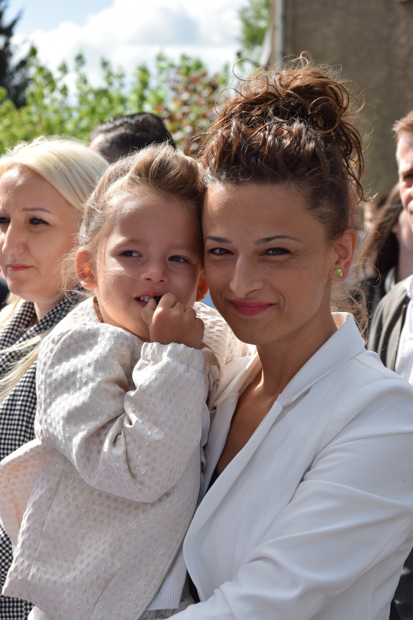 Portret uśmiechniętej kobiety w białym żakiecie trzymającej na rękach małą dziewczynkę w kremowej kurtce na tle innych osób.