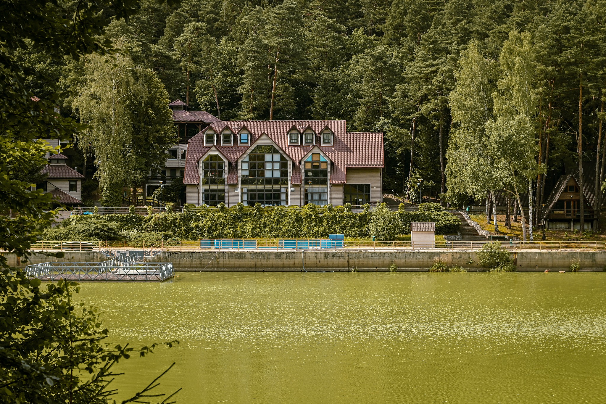 Widok na pensjonat z brązowym dachem i dużymi oknami, otoczony zielenią lasu i stojący nad wodą o oliwkowym zabarwieniu, z widocznym pomostem.