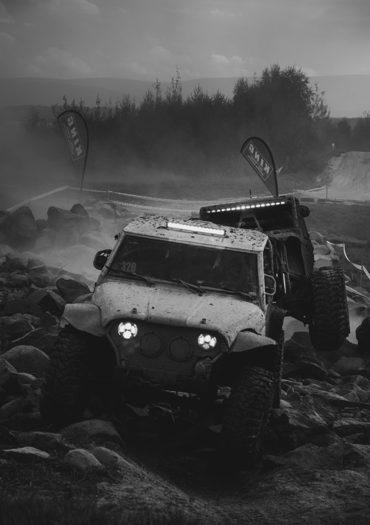 Czarno-białe zdjęcie dwóch samochodów terenowych z numerami startowymi, pokonujących skalisty teren podczas rajdu, z widocznymi flagami i kurzem.