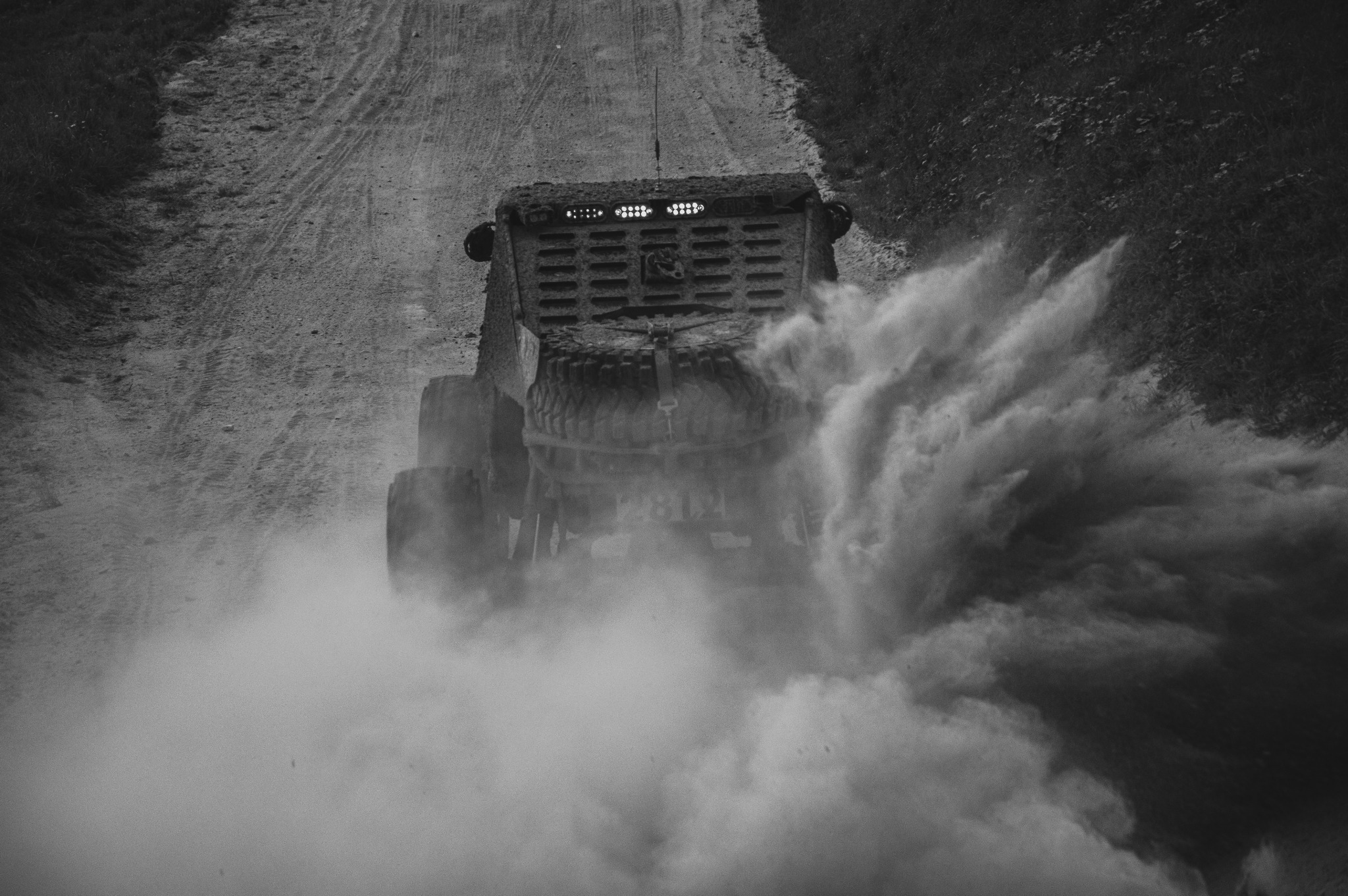 Czarno-białe zdjęcie z tyłu terenowego pojazdu rajdowego w trakcie jazdy po szutrowej drodze, wzbijającego tumany kurzu.