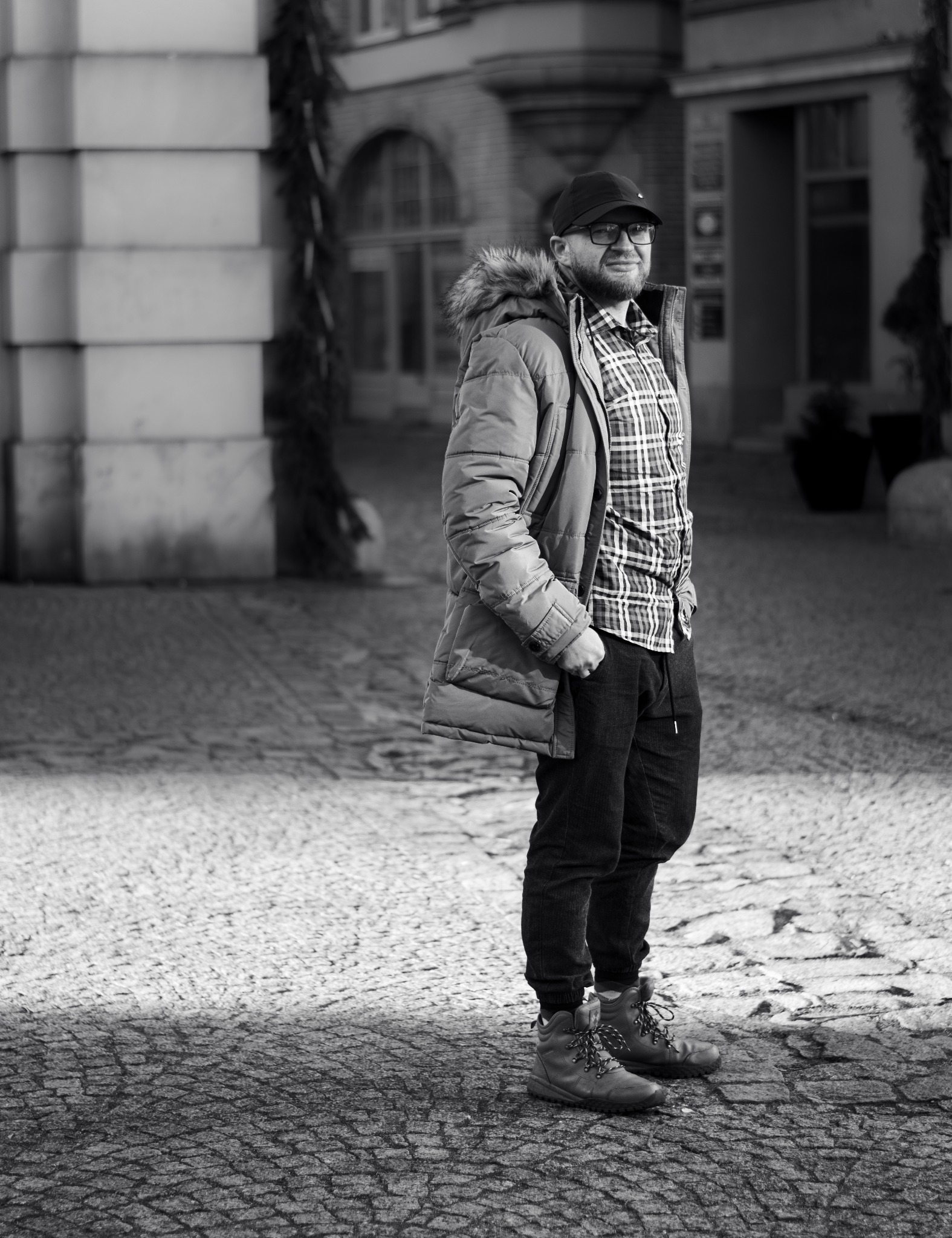 Mężczyzna w zimowej kurtce z kapturem obszytym futrem, czapce i okularach stoi na ulicy z kostki brukowej w czarno-białym ujęciu.