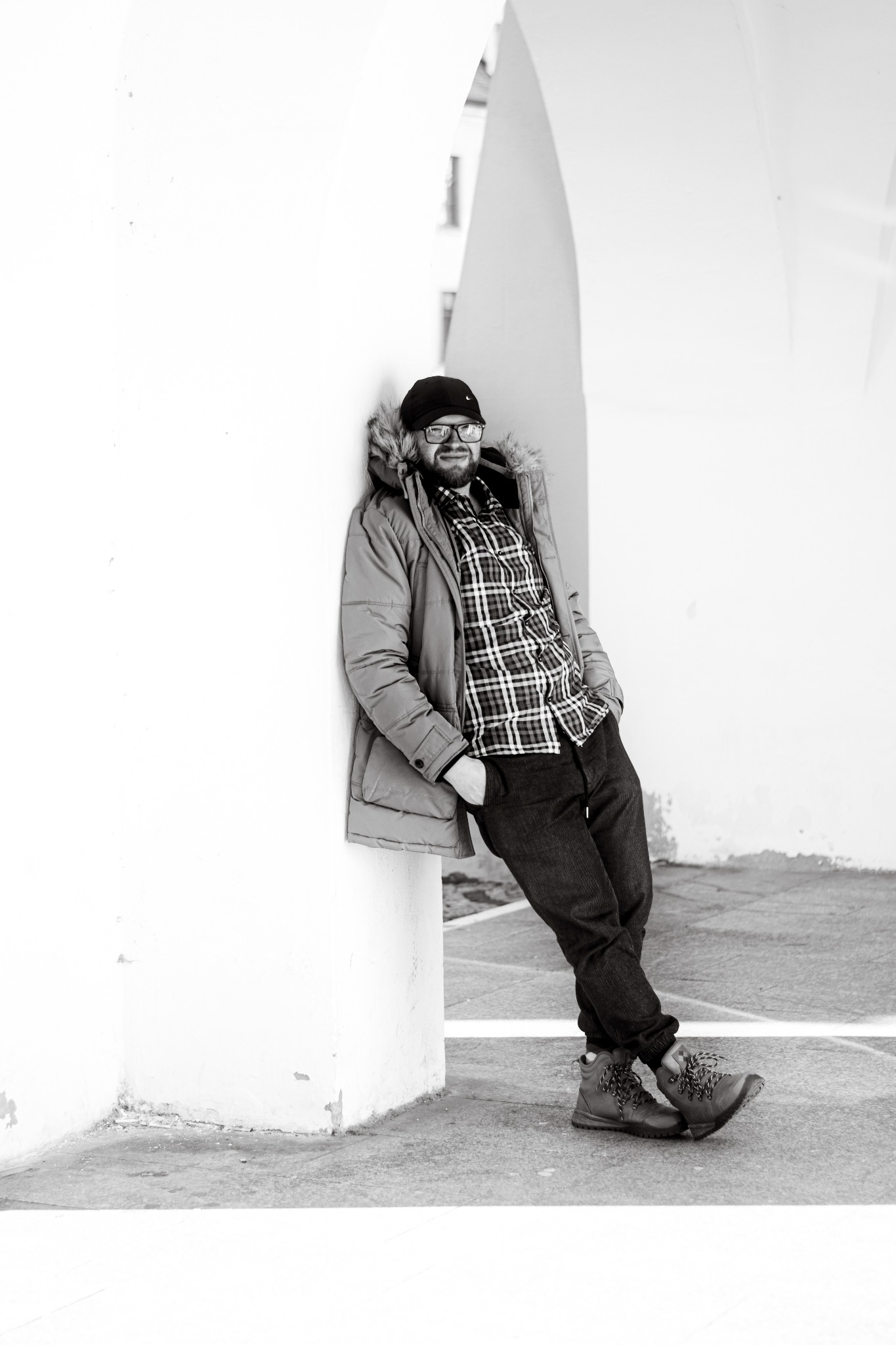 Mężczyzna w okularach, czapce i zimowej kurtce oparty o biały filar w plenerze miejskim, ubrany w koszulę w kratę i ciemne spodnie, pozuje do zdjęcia w czerni i bieli.