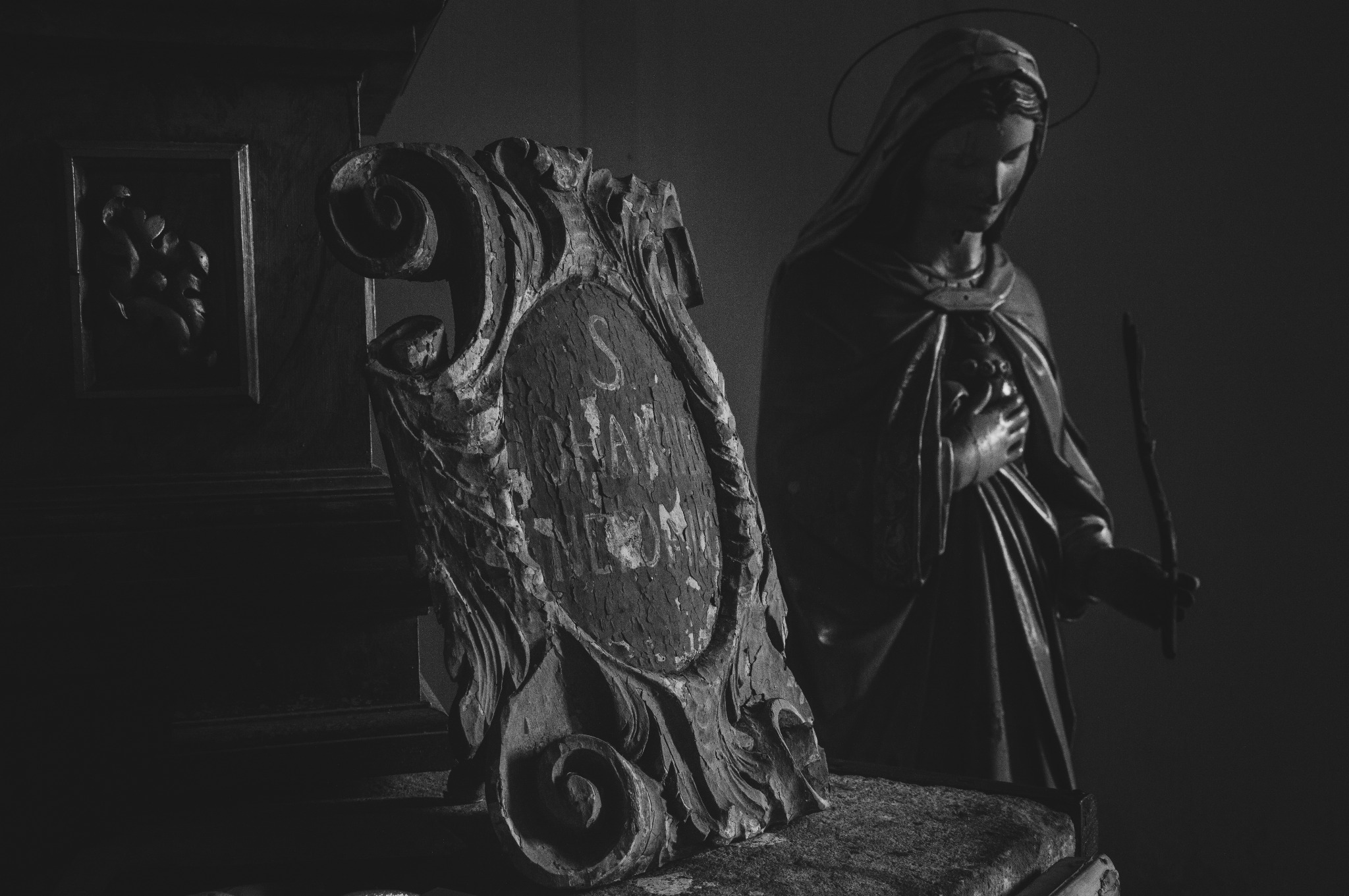 Czarno-białe ujęcie sakralnej rzeźby Matki Boskiej z aureolą, stojącej obok zdobionej, drewnianej tablicy z inskrypcjami w kościelnym wnętrzu.