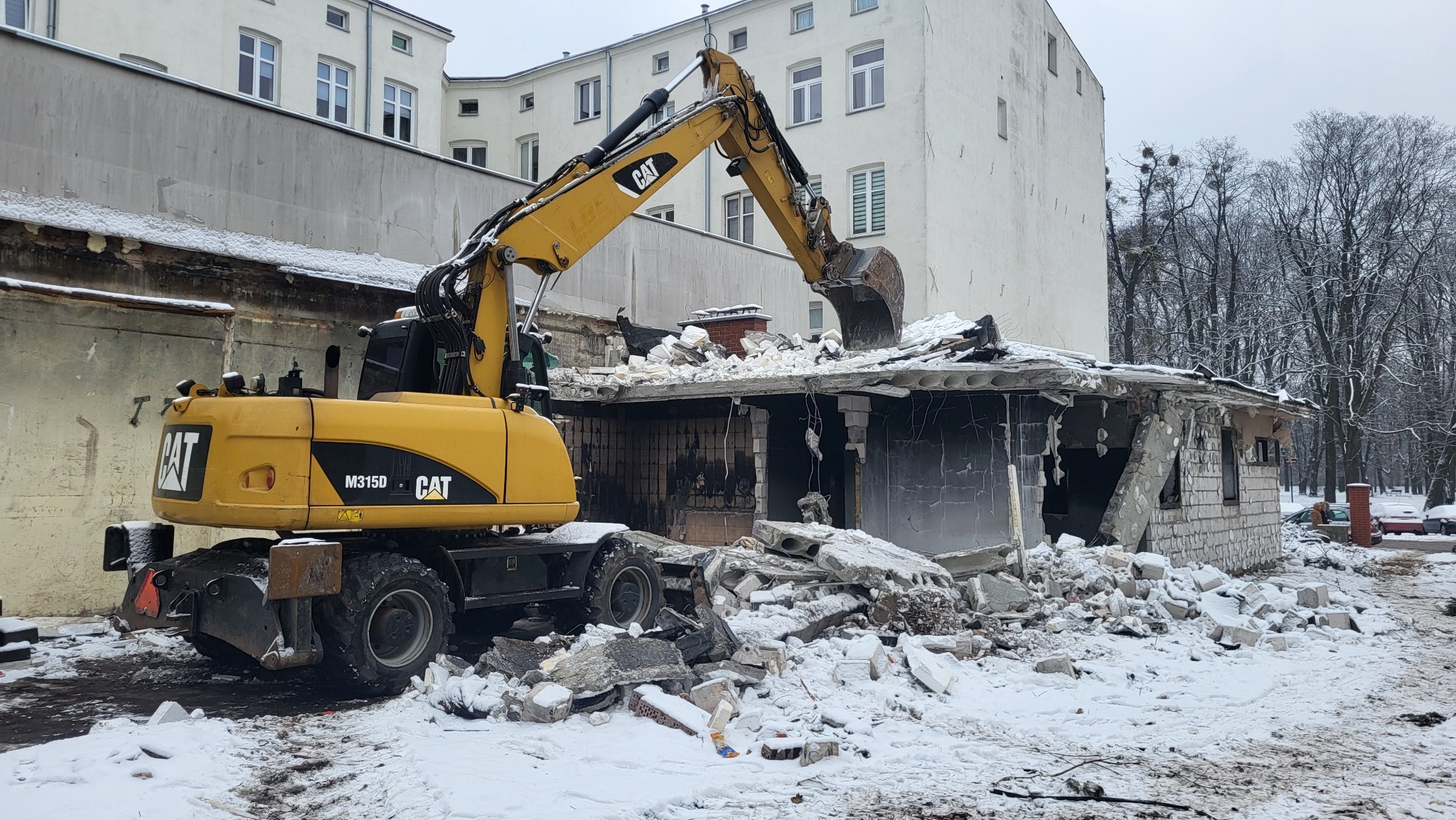 Żółta koparka CAT M315D w trakcie rozbiórki budynku w zimowej scenerii. Gruz i śnieg na pierwszym planie, w tle budynek mieszkalny i drzewa.