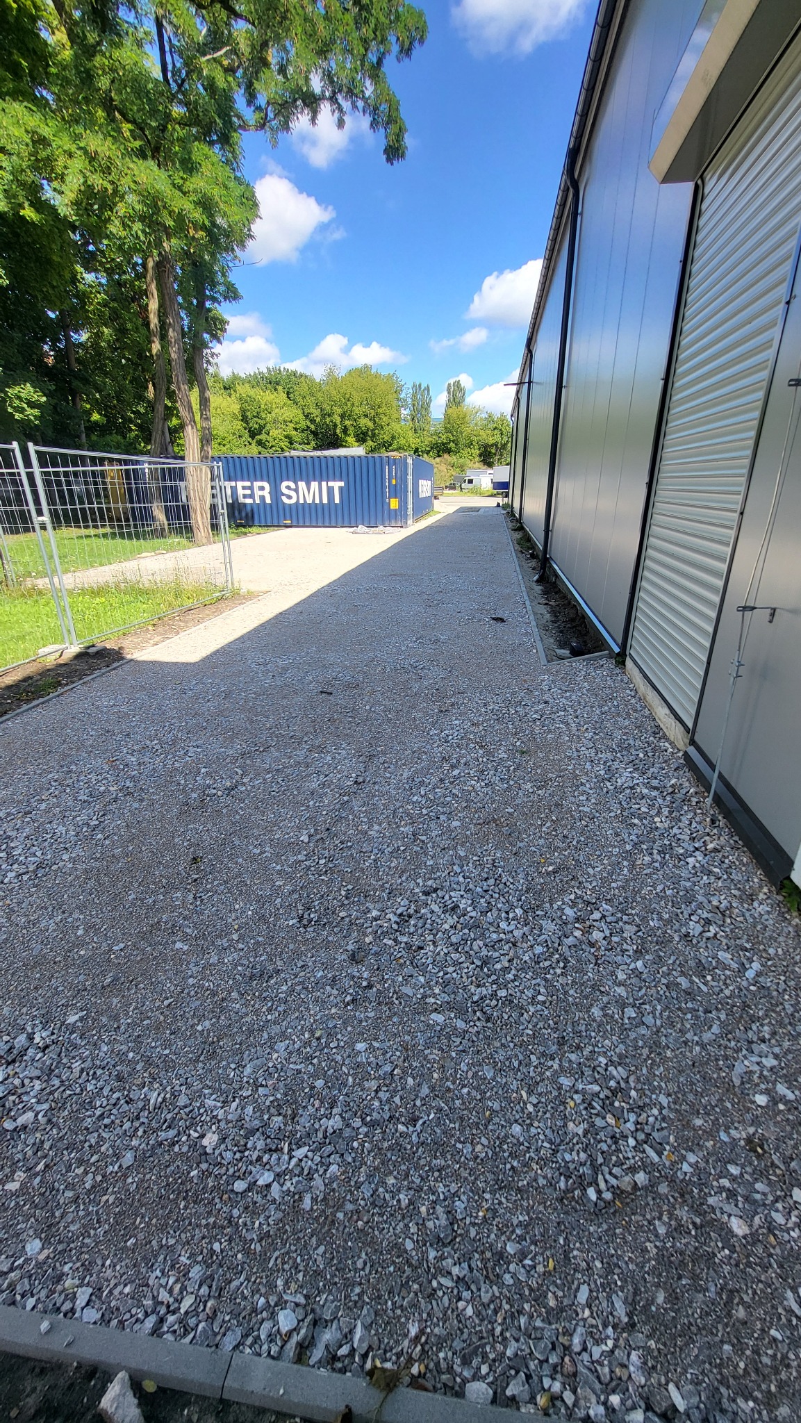 Utwardzona żwirowa droga dojazdowa obok budynku z blachy falistej, w tle kontener i zieleń. Słoneczny dzień, błękitne niebo z chmurami.