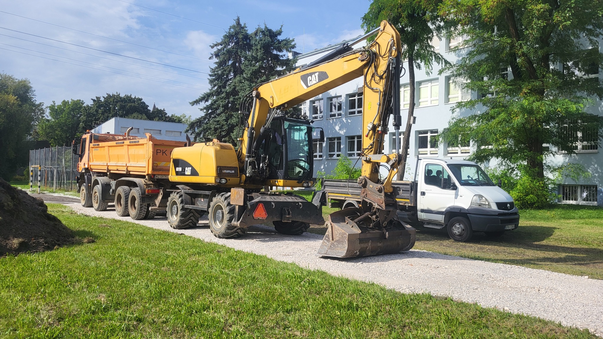 Żółta koparka CAT na tle budynku i wywrotki, na żwirowej drodze. Widoczny fragment trawnika i hałda ziemi. Słoneczny dzień, błękitne niebo.