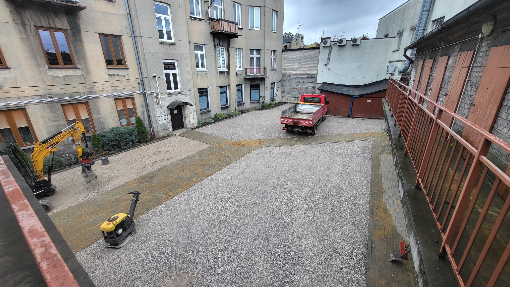 Utwardzony teren żwirowy z mini koparką i zagęszczarką w tle zabudowy miejskiej. Czerwona ciężarówka stoi na nowej nawierzchni. Widok z góry.