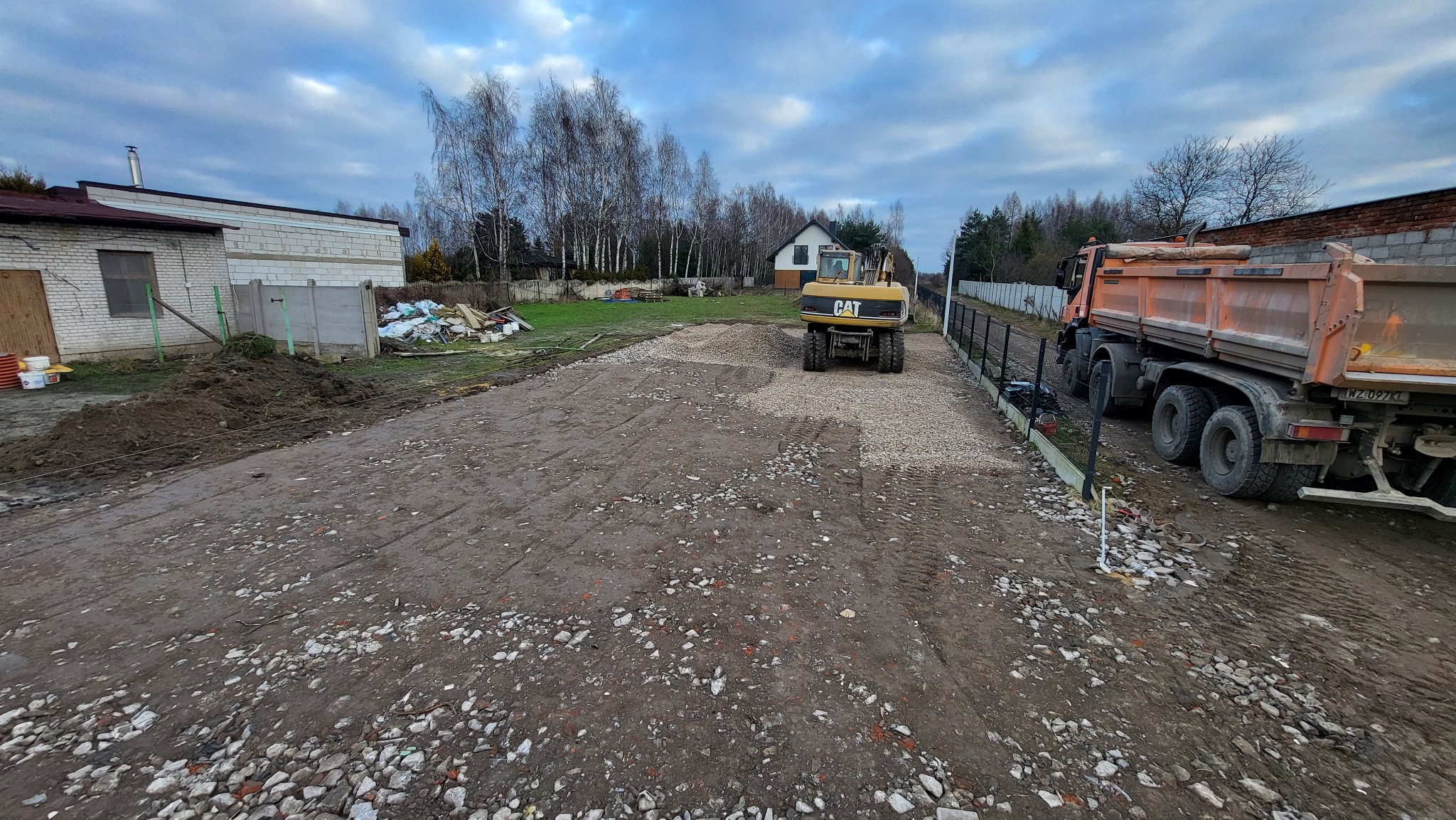 Żółta koparka CAT na utwardzonym żwirem terenie budowy, obok pomarańczowa wywrotka. W tle budynki i drzewa pod pochmurnym niebem.