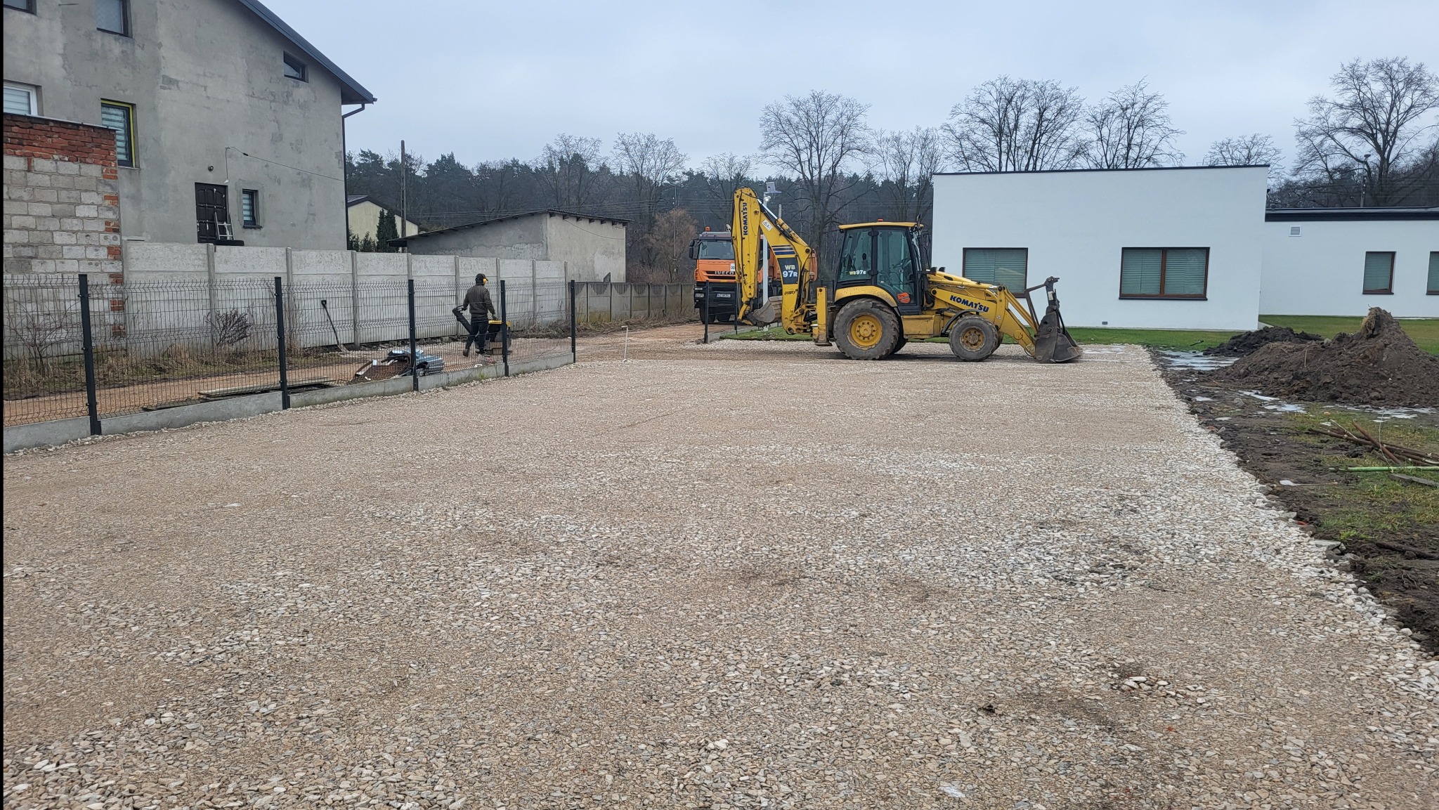 Żwirowa nawierzchnia wyrównana koparko-ładowarką Komatsu WB97S, w tle ogrodzenie panelowe i budynki. Prace ziemne na działce.