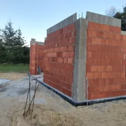 Wznoszone ściany budynku z czerwonej cegły ceramicznej, z widoczną izolacją przeciwwilgociową fundamentów i częściowo wylanymi betonowymi nadprożami, na tle piaszczystego terenu budowy i drzew.