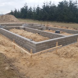 Rafał Stępiński - Wznoszenie fundamentów z szarych bloczków betonowych na piaszczystym terenie; widoczny szalunek z drewnianych desek i sterczące pręty zbrojeniowe.