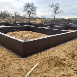 Rafał Stępiński - Zabezpieczone, pomalowane na ciemno, fundamenty domu jednorodzinnego z widoczną konstrukcją szalunkową, piaskiem w środku i rurą kanalizacyjną, na piaszczystym terenie, w tle drzewa bez liści...