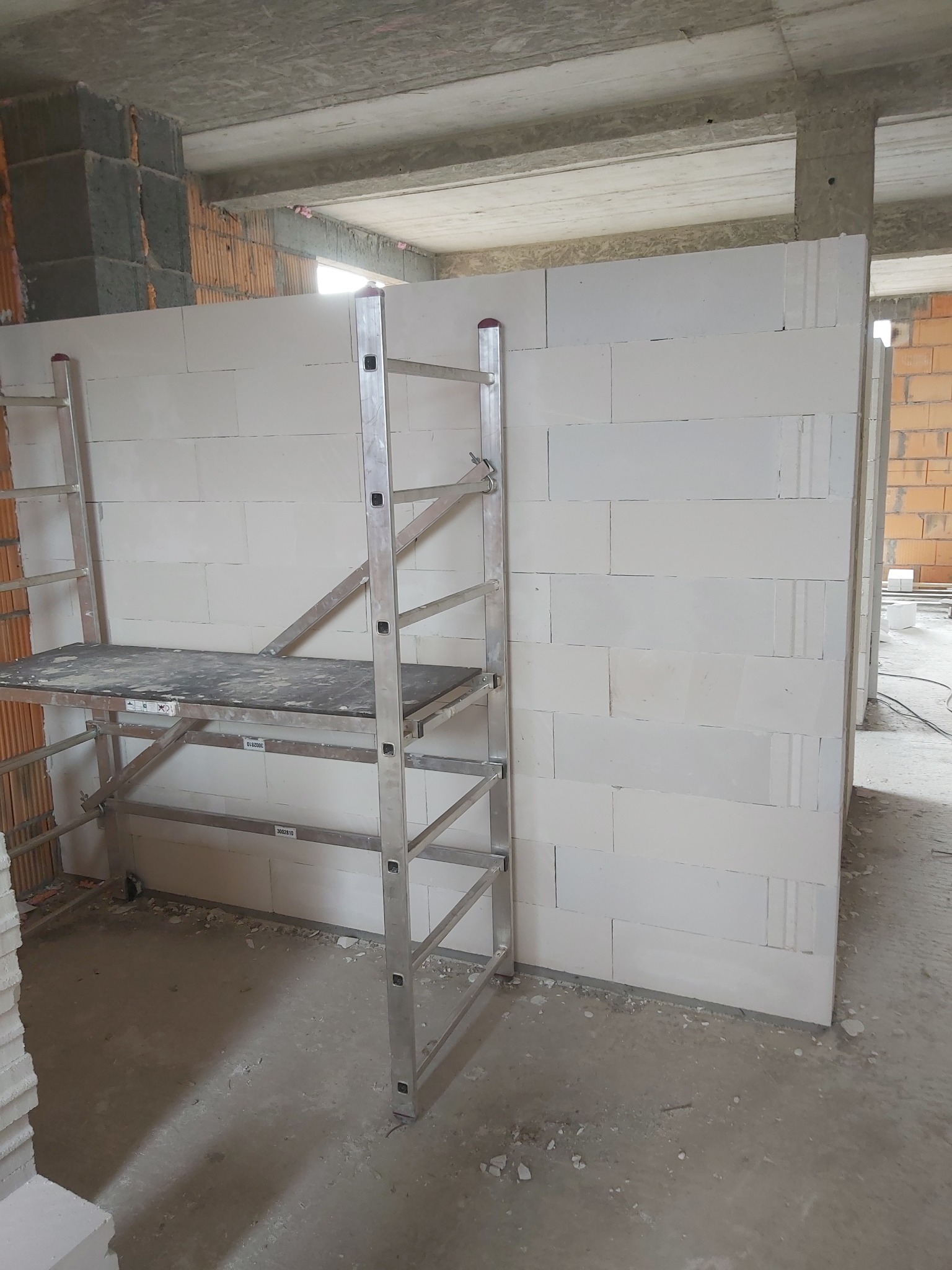 Wzniesiona ściana z białych bloczków w surowym stanie, widoczne rusztowanie aluminiowe z drewnianym podestem, betonowy strop w tle, ślady zaprawy na podłodze.