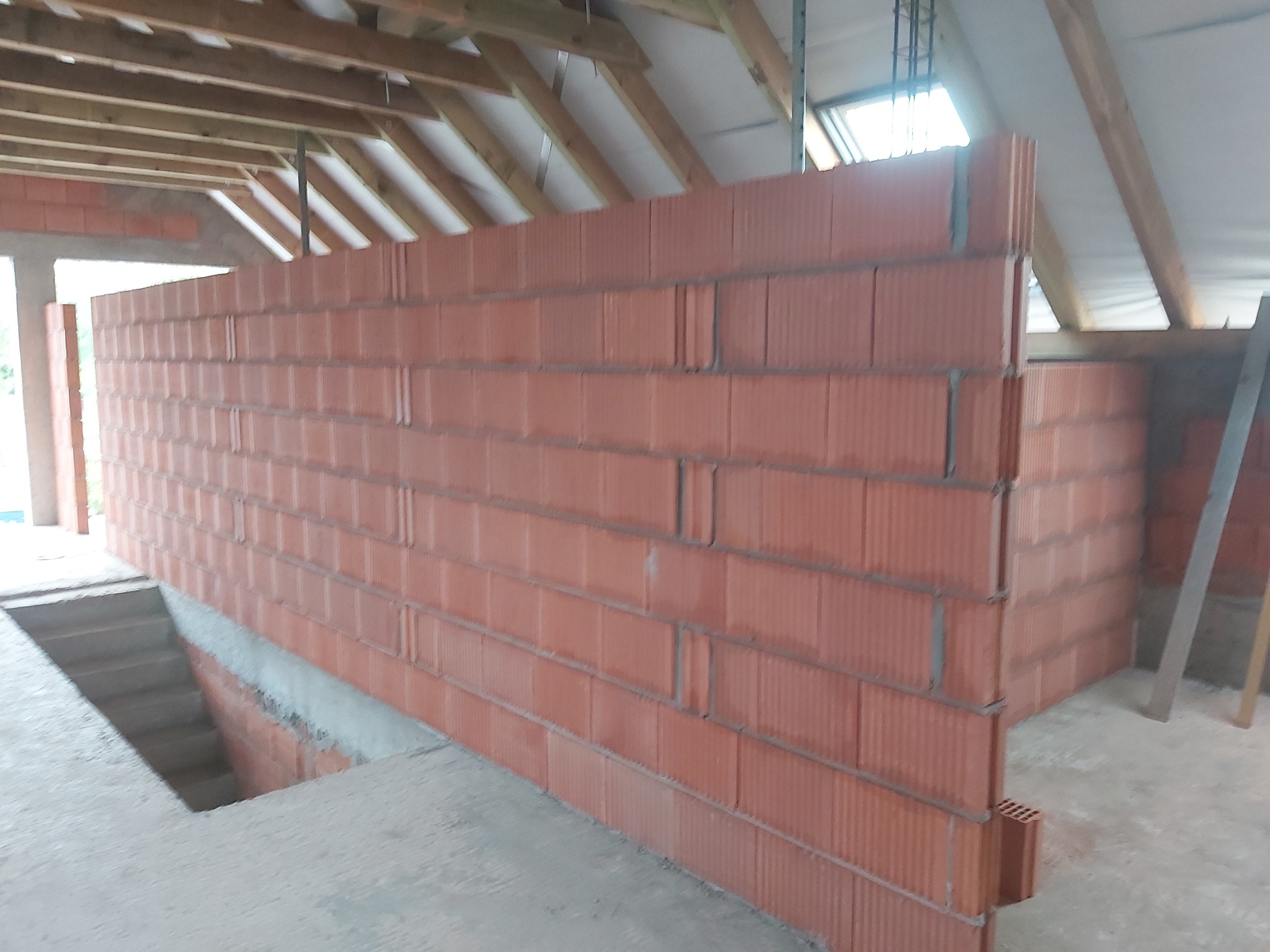 Ściana z czerwonych bloczków ceramicznych w surowym stanie, widoczna konstrukcja drewniana dachu i betonowa podłoga w trakcie budowy poddasza.