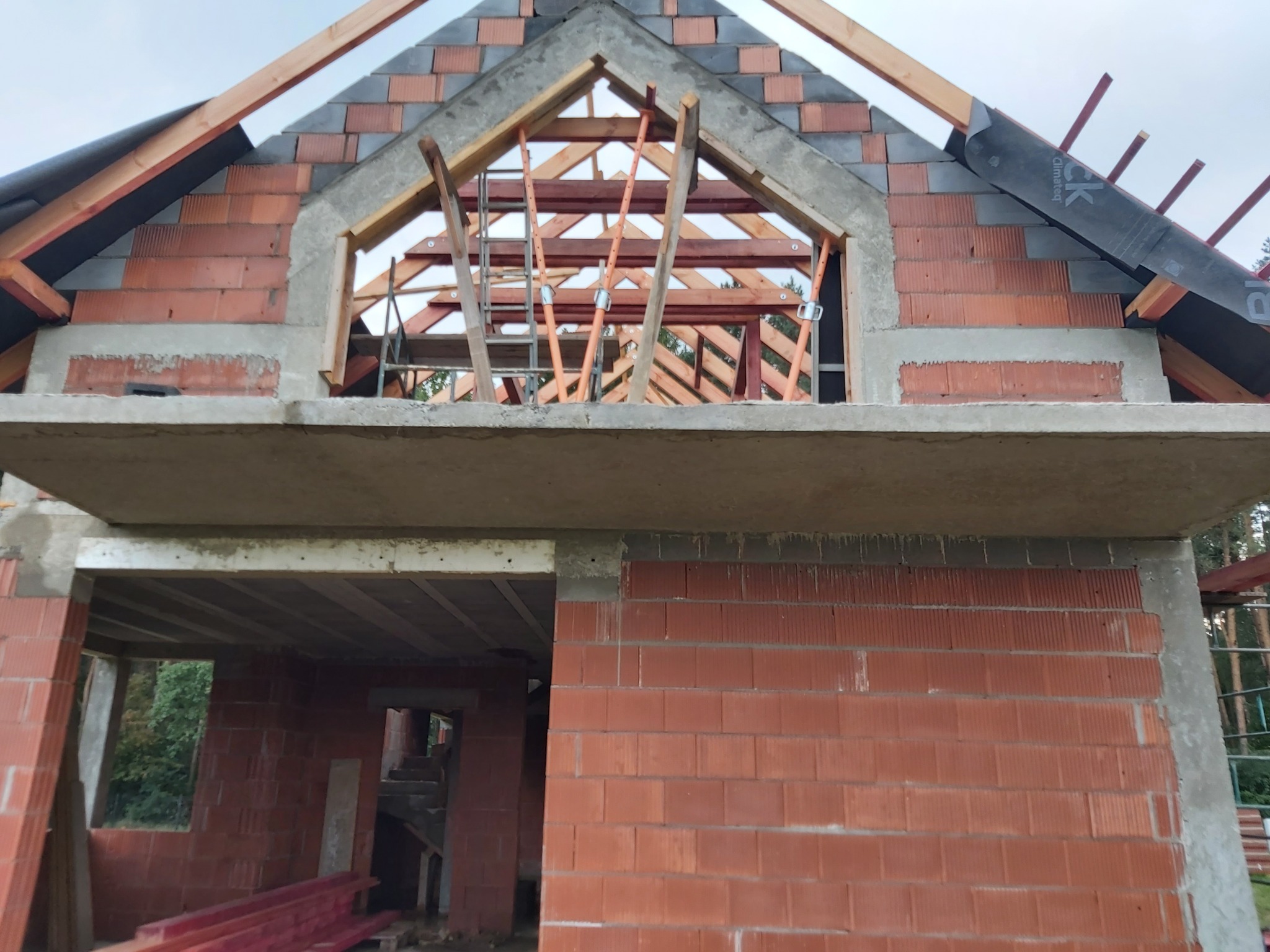 Surowy stan budynku z czerwonej cegły ceramicznej, widoczne drewniane elementy konstrukcji dachu i betonowy strop, w tle zieleń drzew.