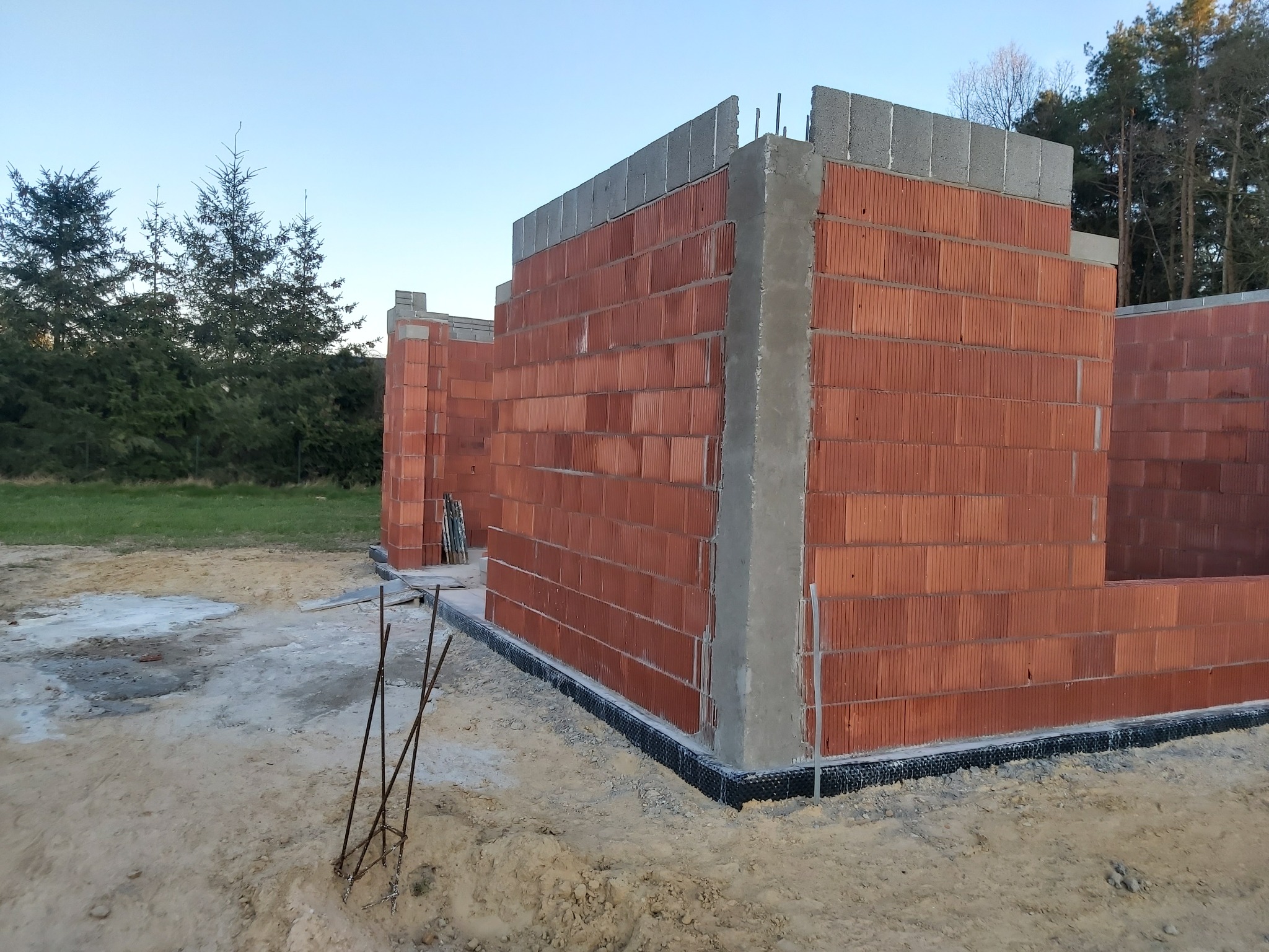Wznoszone ściany budynku z czerwonej cegły ceramicznej, z widoczną izolacją przeciwwilgociową fundamentów i częściowo wylanymi betonowymi nadprożami, na tle piaszczystego terenu budowy i drzew.