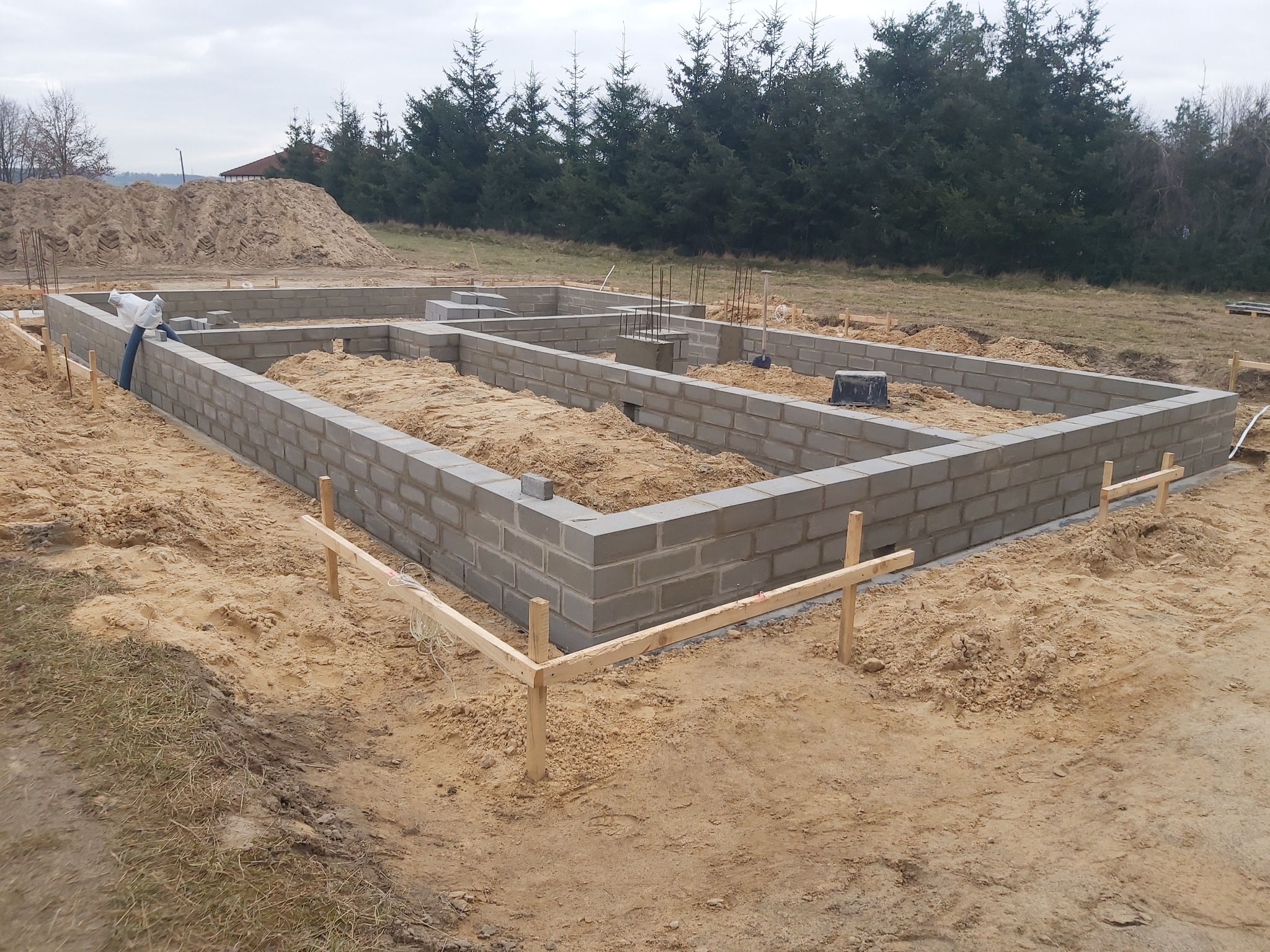 Wznoszenie fundamentów z szarych bloczków betonowych na piaszczystym terenie; widoczny szalunek z drewnianych desek i sterczące pręty zbrojeniowe.