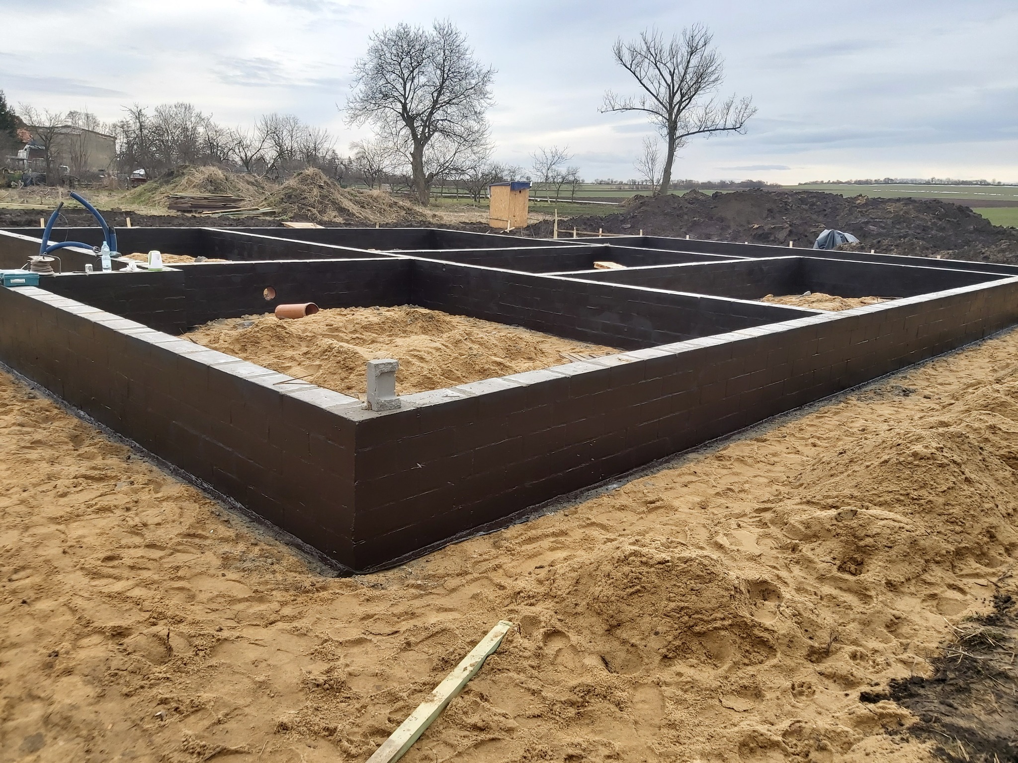 Zabezpieczone, pomalowane na ciemno, fundamenty domu jednorodzinnego z widoczną konstrukcją szalunkową, piaskiem w środku i rurą kanalizacyjną, na piaszczystym terenie, w tle drzewa bez liści...