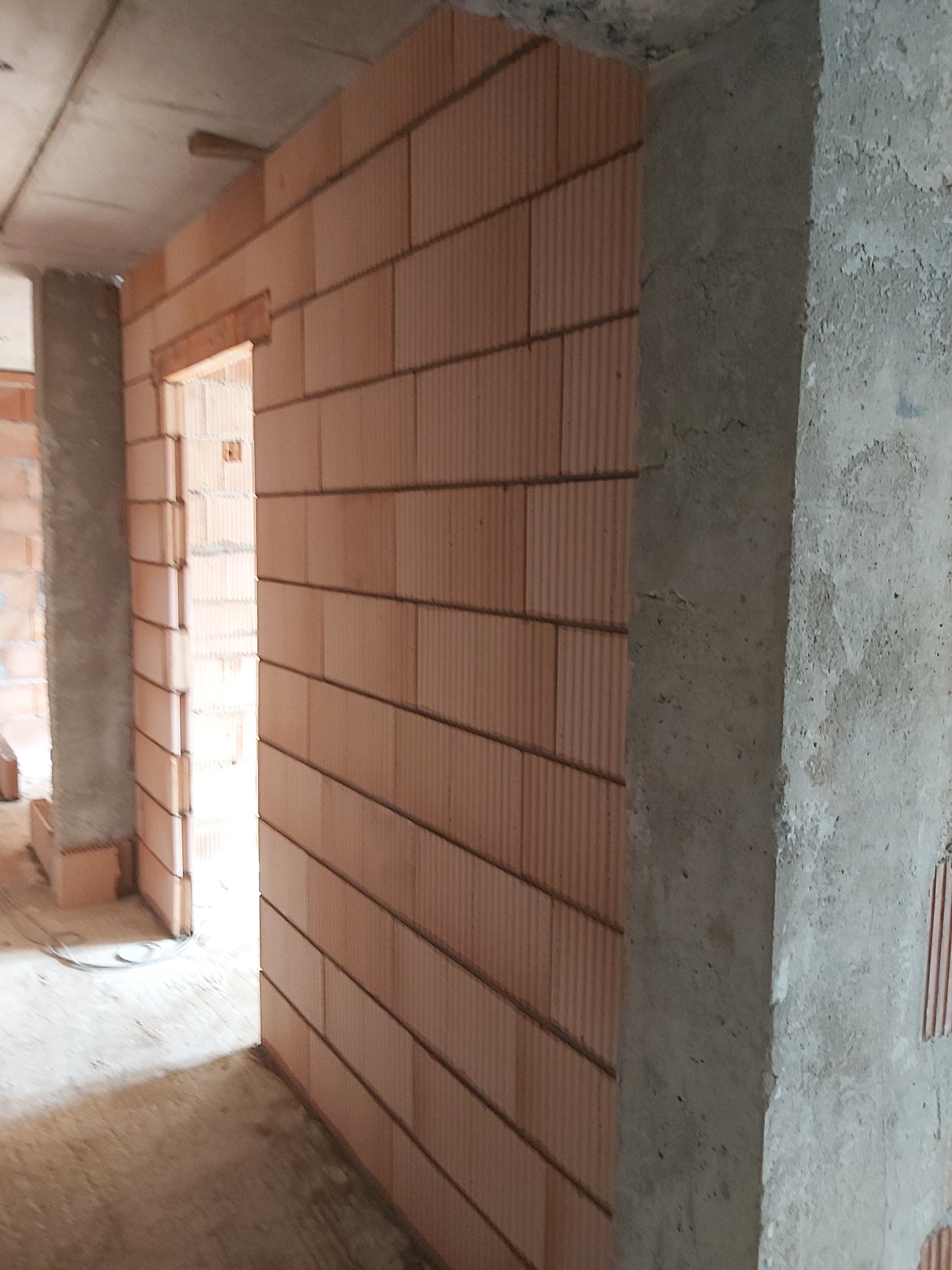 Wznoszenie ściany z pustaków ceramicznych w surowym wnętrzu, widoczny fragment otworu drzwiowego i surowy betonowy filar.