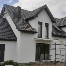 Nowoczesny dom jednorodzinny z białą elewacją i grafitowym dachem z blachodachówki, fragment dachu z odkrytą konstrukcją drewnianą, rusztowanie przy oknie balkonowym, widoczne ślady po pracach...