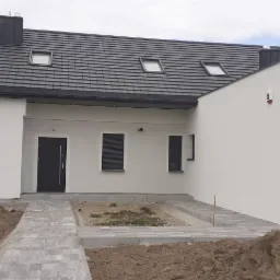 Nowoczesny dom jednorodzinny z szarą dachówką i białymi ścianami, widoczne prace ziemne i układanie kostki brukowej przed budynkiem, pracownik w tle.