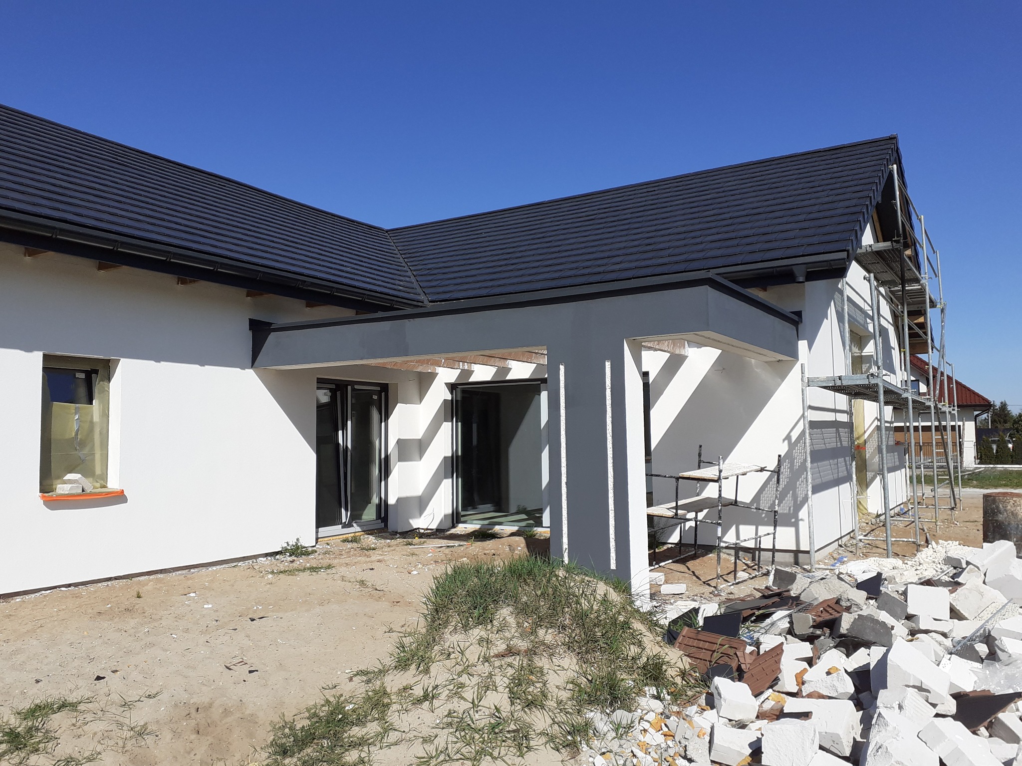 Nowo otynkowana, biała elewacja domu jednorodzinnego z czarną dachówką, z widocznymi rusztowaniami i stertą gruzu po pracach wykończeniowych, w słoneczny dzień.