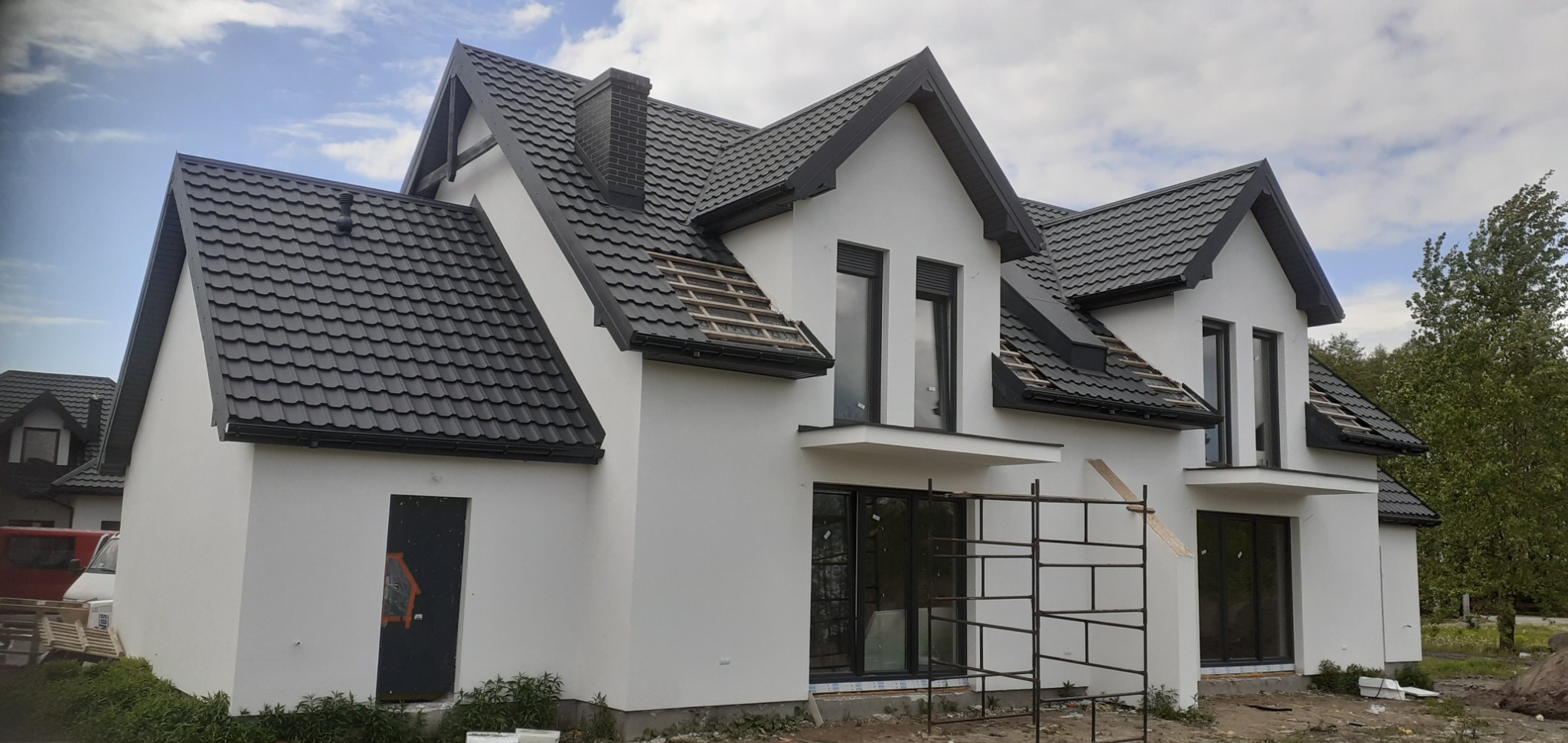 Nowoczesny dom jednorodzinny z białą elewacją i grafitowym dachem z blachodachówki, fragment dachu z odkrytą konstrukcją drewnianą, rusztowanie przy oknie balkonowym, widoczne ślady po pracach...