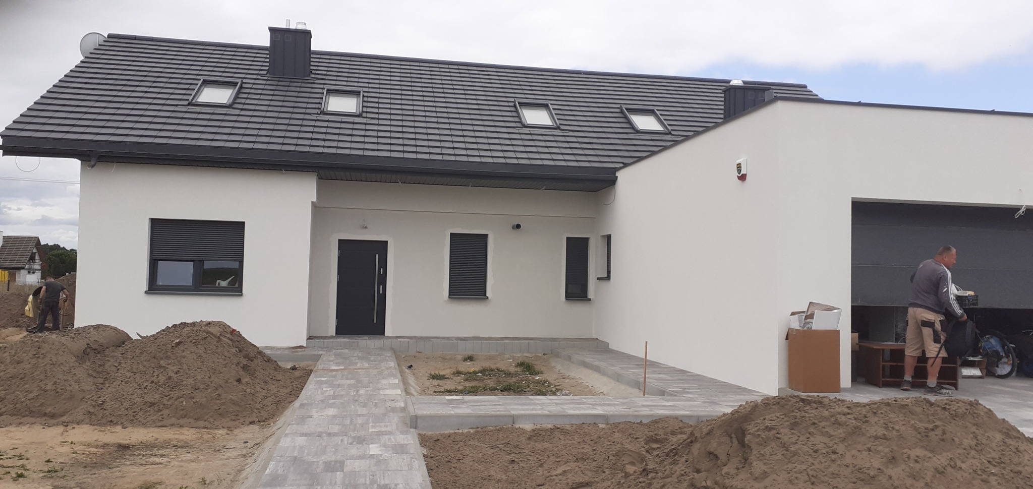 Nowoczesny dom jednorodzinny z szarą dachówką i białymi ścianami, widoczne prace ziemne i układanie kostki brukowej przed budynkiem, pracownik w tle.