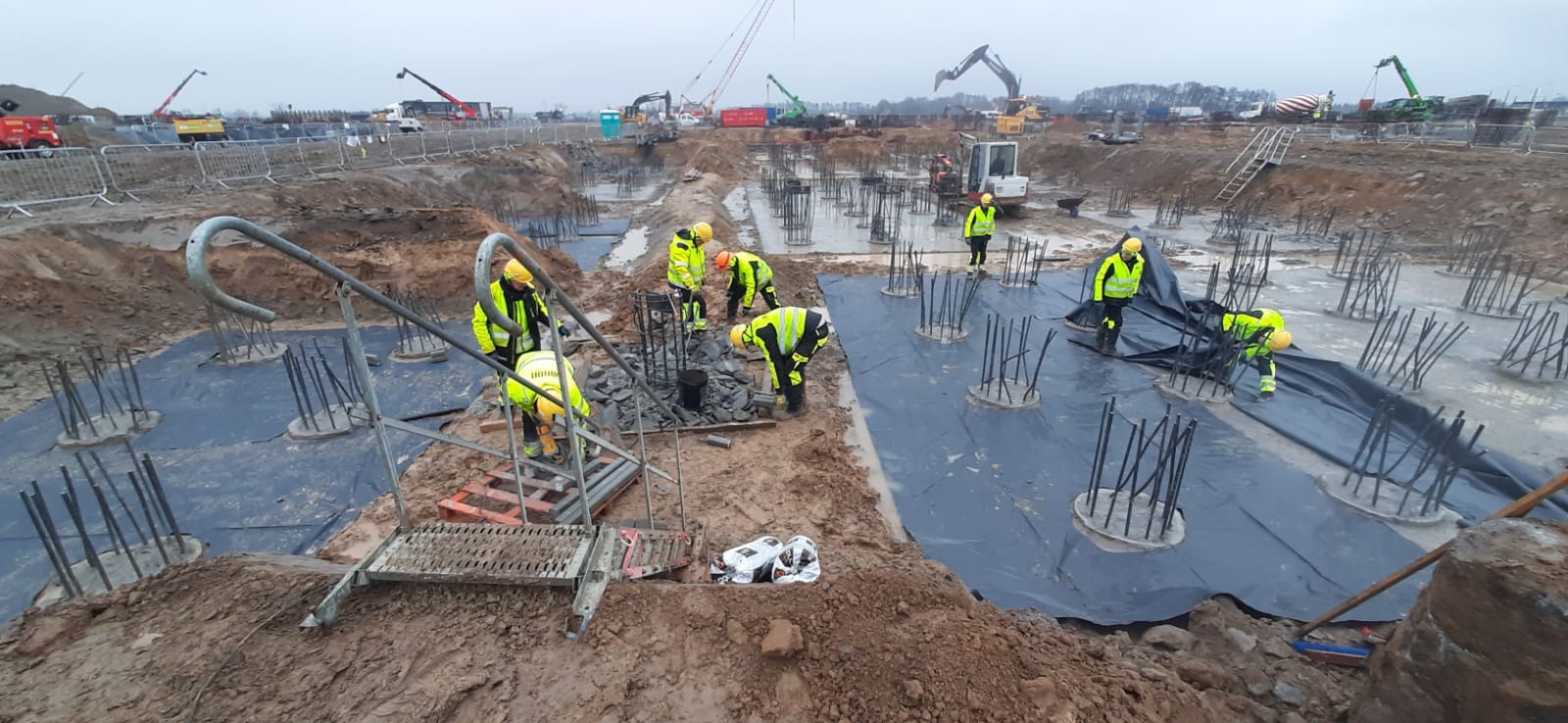 Prace ziemne na placu budowy z widocznymi fundamentami pod budynek żelbetowy, zbrojeniami i pracownikami w odblaskowych kamizelkach. W tle sprzęt budowlany i pochmurne niebo.