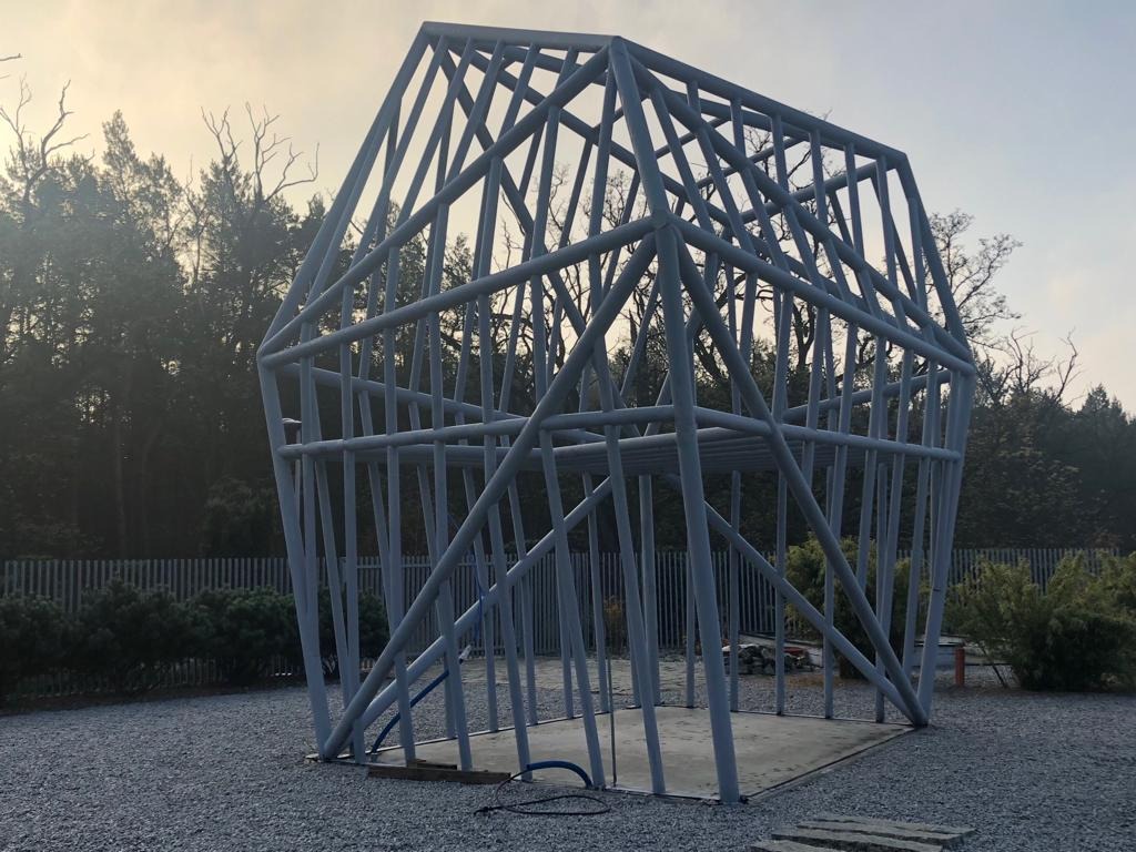 Szkielet budynku o nietypowym kształcie z metalowych rur na betonowej płycie, otoczony szarym żwirem i zielenią, widok z zewnątrz w pochmurny dzień.