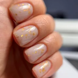 Delikatny, mleczny manicure hybrydowy z subtelnymi złotymi płatkami na krótkich, kwadratowych paznokciach.