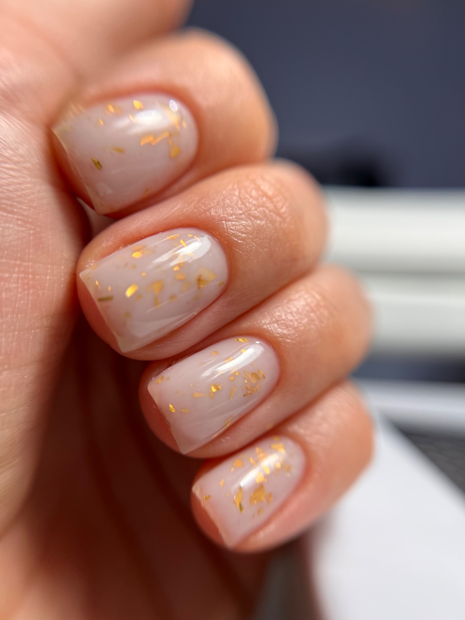 Delikatny, mleczny manicure hybrydowy z subtelnymi złotymi płatkami na krótkich, kwadratowych paznokciach.
