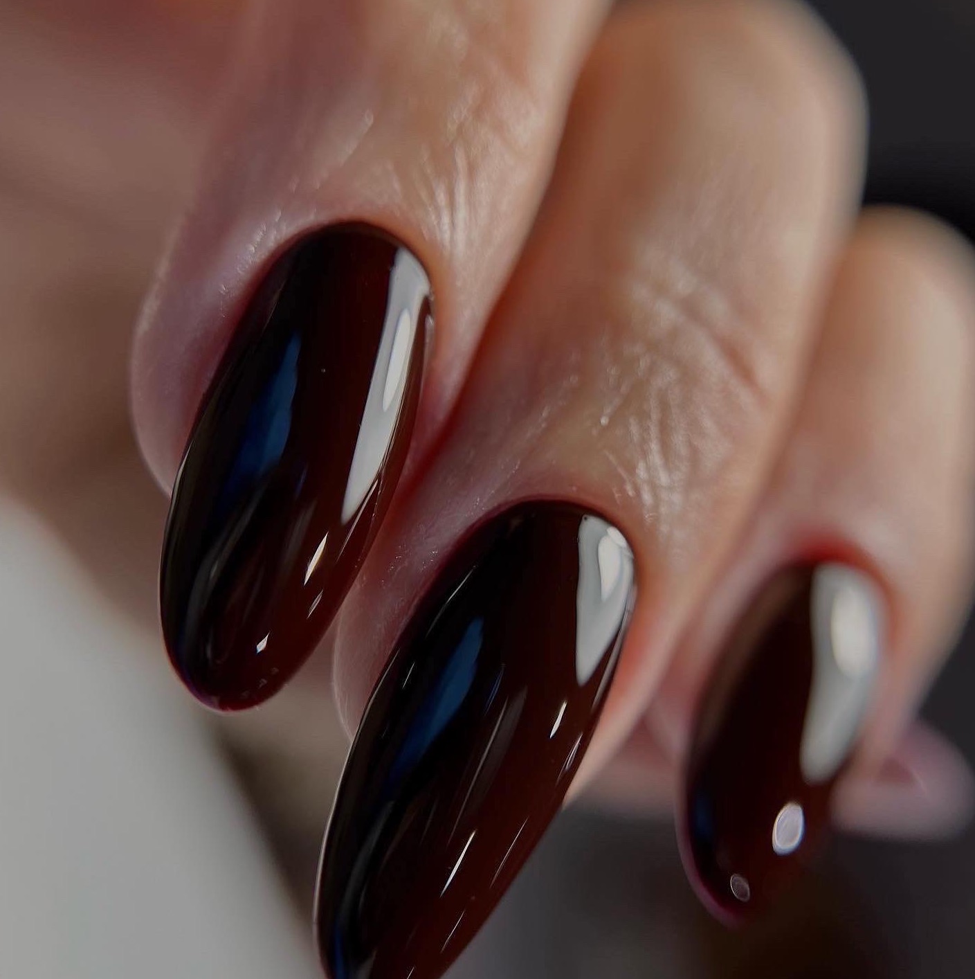 Błyszczące, ciemnowiśniowe paznokcie o owalnym kształcie, z widocznymi refleksami światła, na tle skóry dłoni.