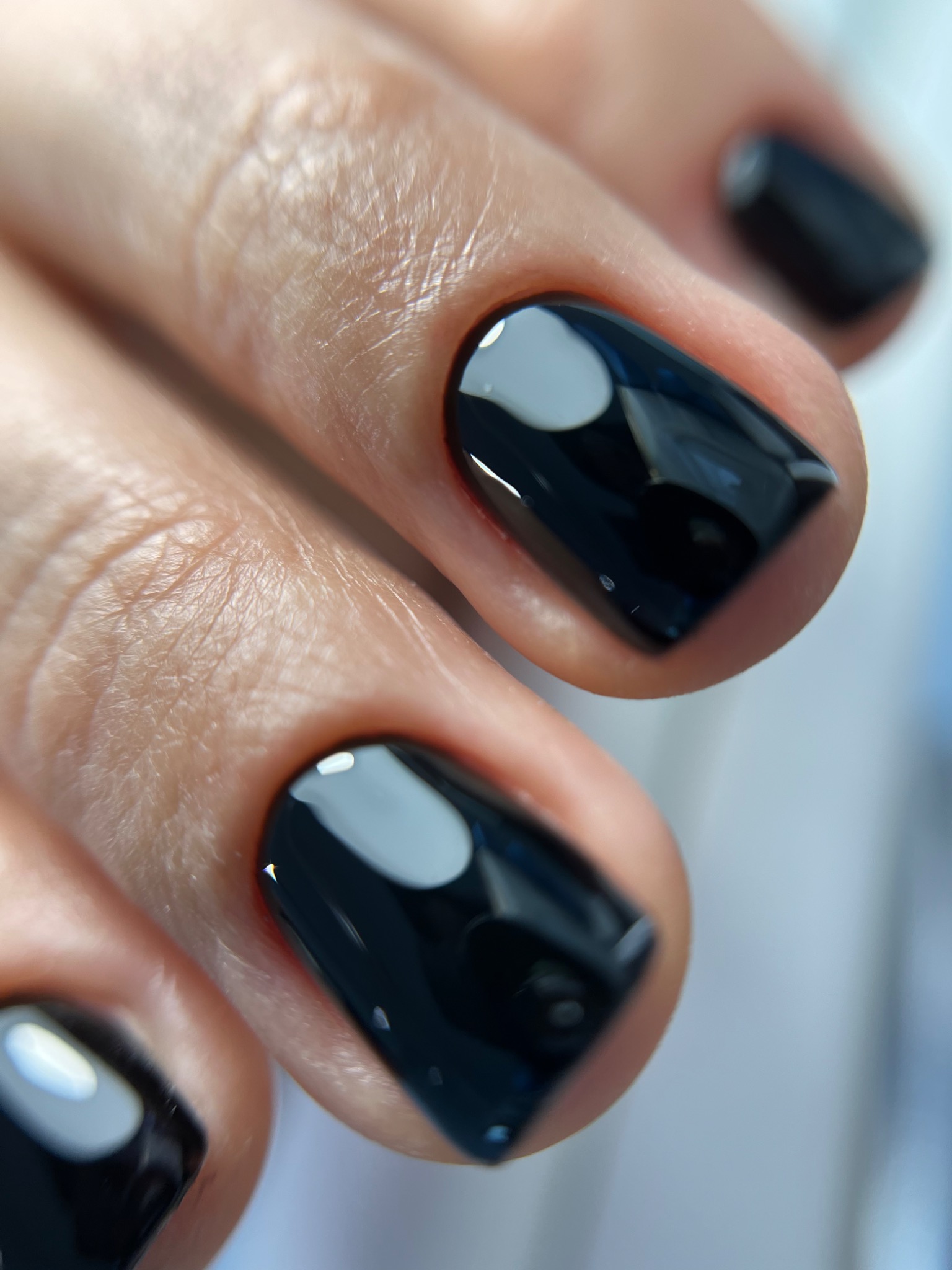 Zbliżenie na dłoń z krótkimi, kwadratowymi paznokciami pomalowanymi na błyszczący, ciemny granat, z widocznymi refleksami światła.