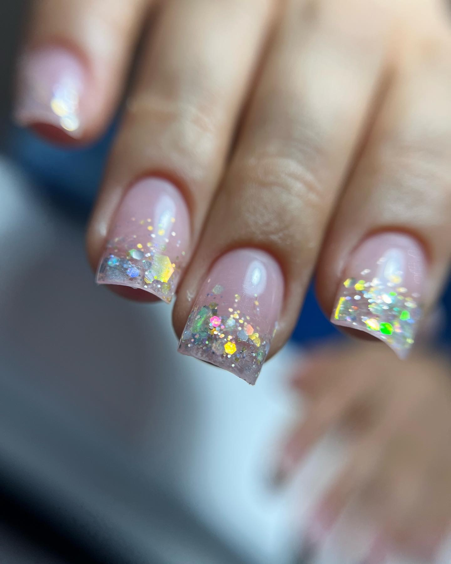 Zbliżenie na dłoń z kwadratowymi paznokciami pokrytymi jasnoróżowym lakierem i brokatowym zdobieniem na końcach, z widocznymi wielokolorowymi, holograficznymi drobinkami.