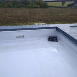 Biała, płaska powierzchnia dachu z odpływem zabezpieczonym kratką, widok z góry, w tle zieleń i wiadukt.