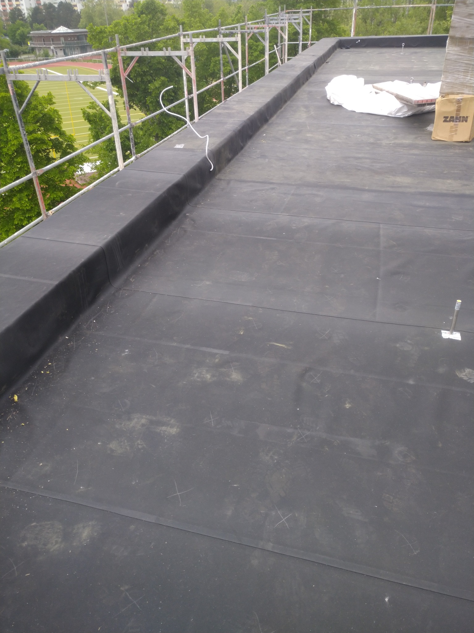 Widok z góry na płaski dach pokryty czarną membraną bitumiczną, zabezpieczony rusztowaniem, z widocznymi śladami stóp i zarysowaniami, z zapasem materiałów budowlanych i kartonem ZAHN w rogu.