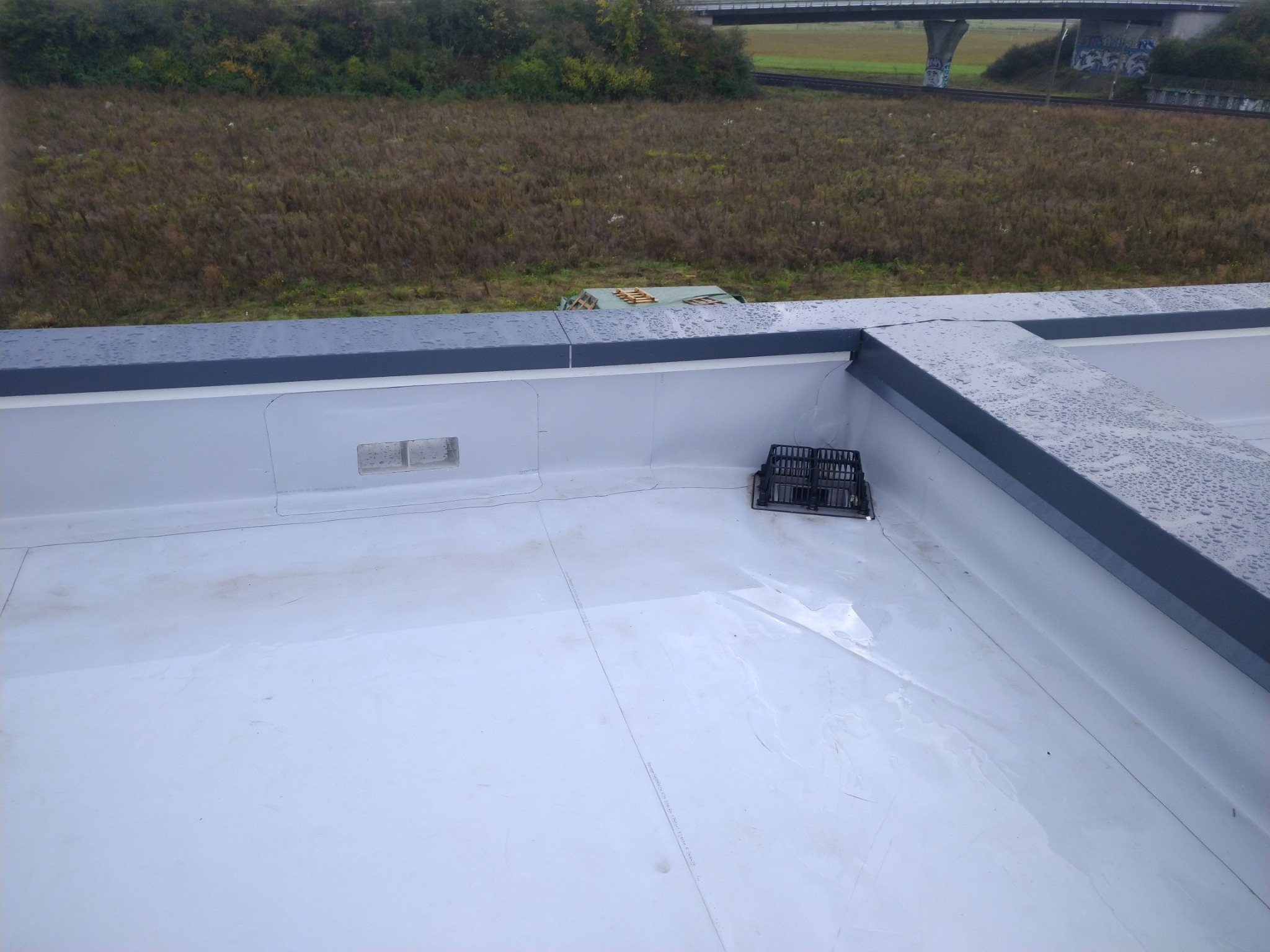 Biała, płaska powierzchnia dachu z odpływem zabezpieczonym kratką, widok z góry, w tle zieleń i wiadukt.