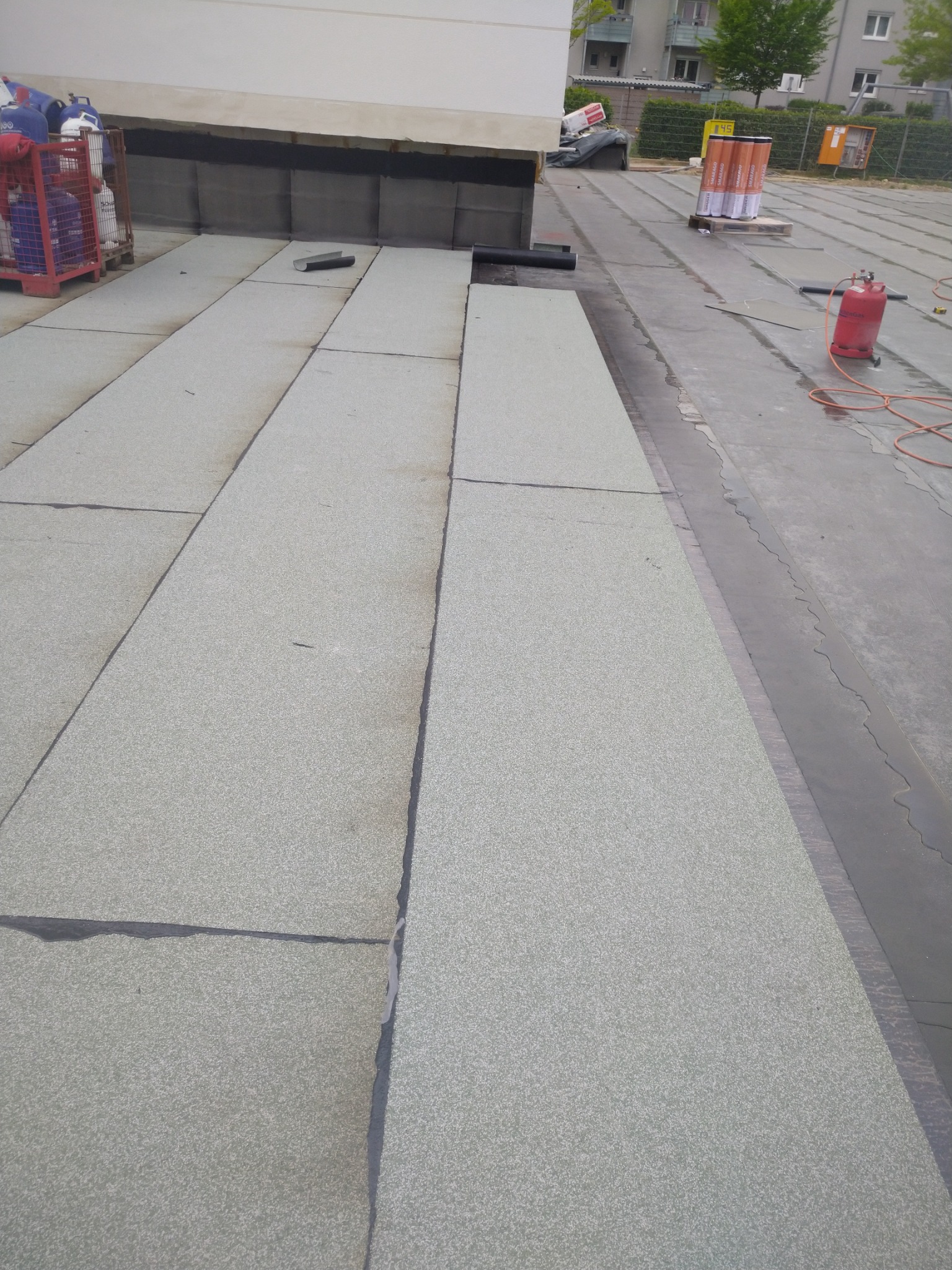 Fragment dachu płaskiego pokrytego papą termozgrzewalną z widocznymi zgrzewami, butla gazowa i rolki papy w tle.